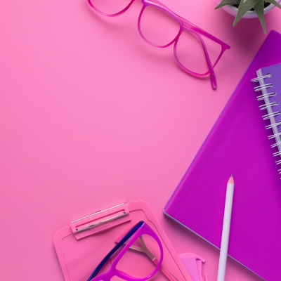 A neatly arranged desk with pink and purple stationery including a notebook, glasses, pens, and a clipboard on a pink background.